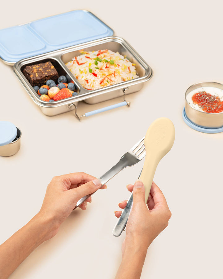 Person holding a fork and spoon with a bento box containing food on a light surface.