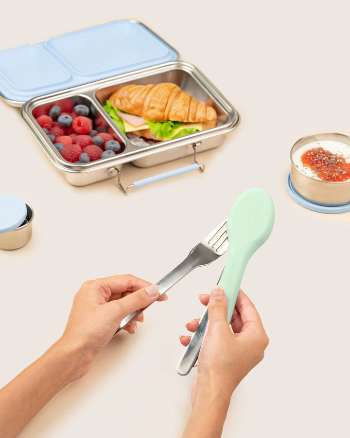Lunchbox with compartments containing food, a fork, and a hand holding stainless steel fork from mint silicone case beside kids’ school tiffin lunchbox India Baboons.