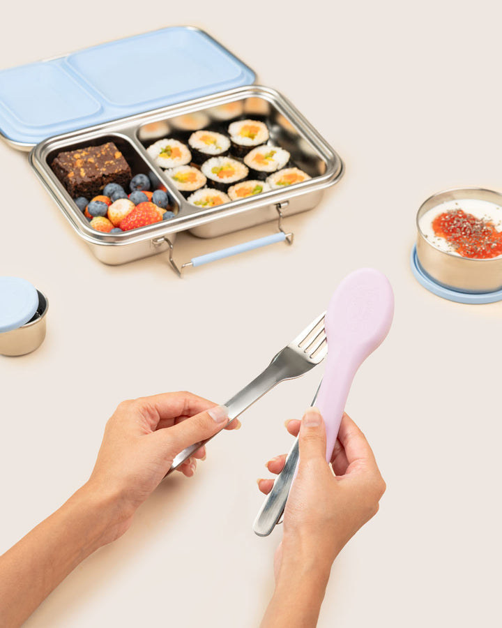 Hands holding stainless steel fork spoon and knife from lilac silicone case beside kids’ steel tiffin box with sushi, fruits in India by Baboons.