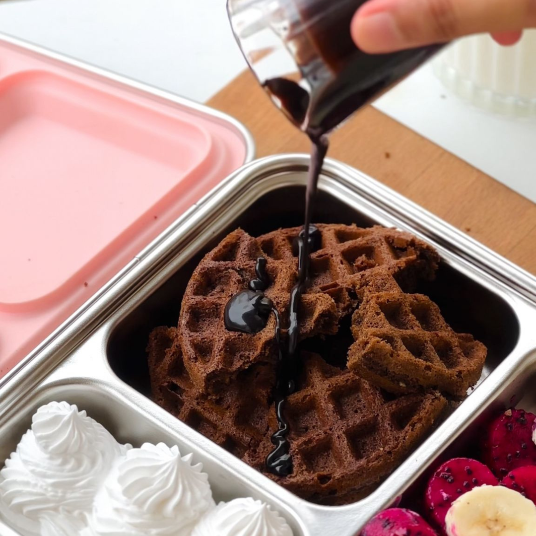 Close-up of chocolate syrup being poured over crispy eggless chocolate waffles packed in a stainless-steel lunchbox, with whipped cream swirls and slices of banana and dragon fruit on the side.