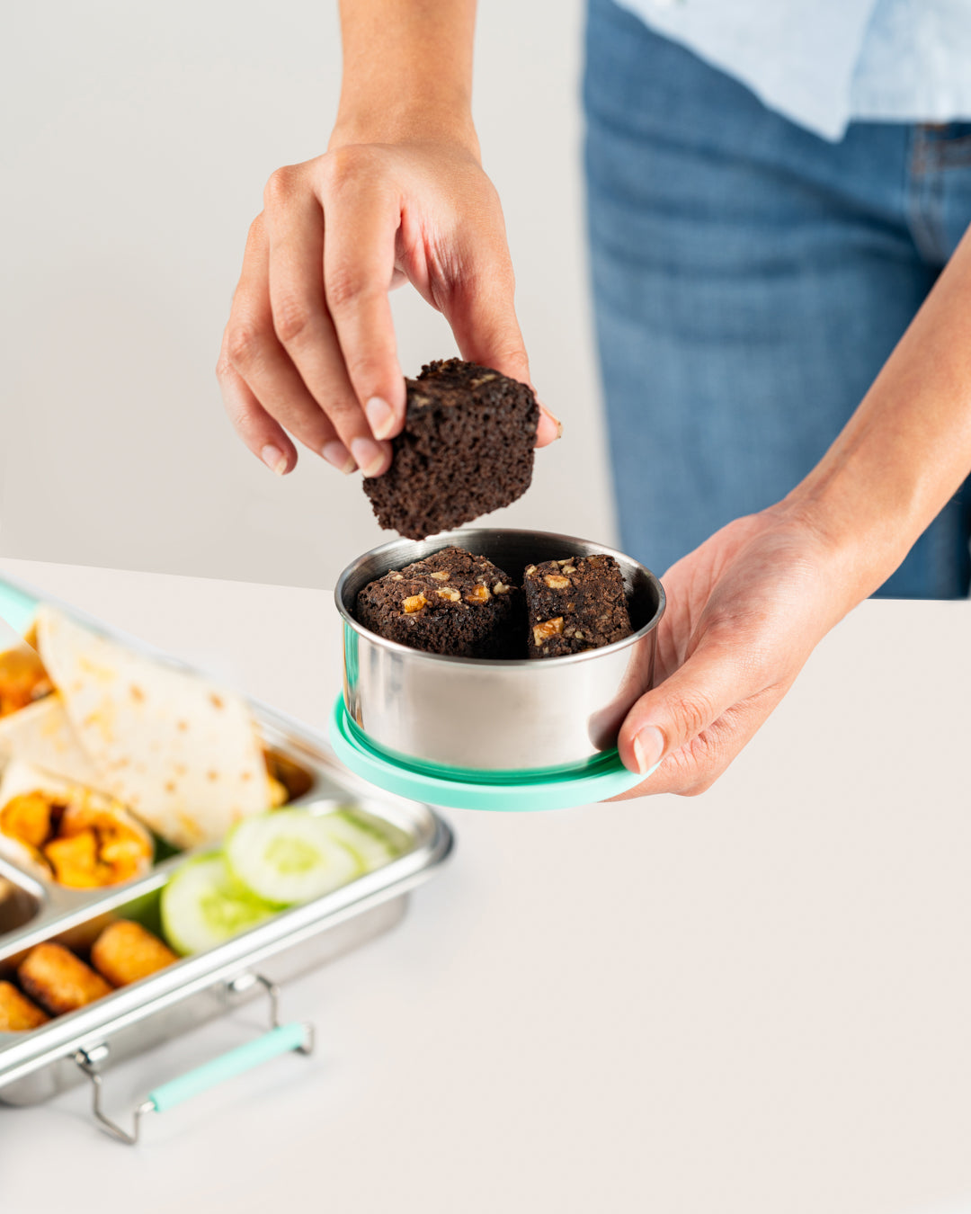 Hand holding stainless steel snack container with aqua silicone lid, filled with brownies, next to a bento lunch box with assorted snacks, perfect for Indian lunch needs