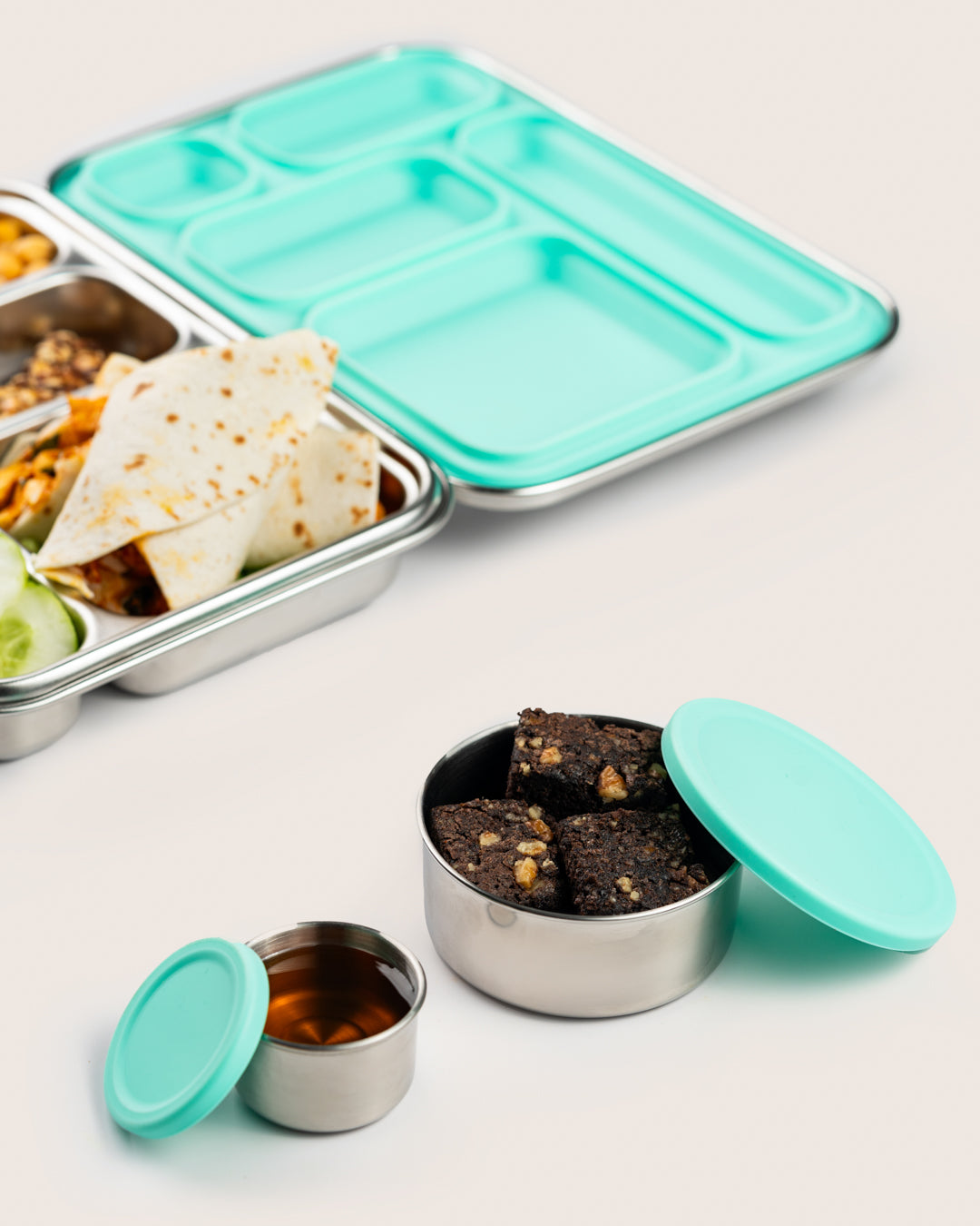 Stainless steel snack containers with aqua silicone lids, filled with brownies and syrup, alongside a bento lunch box, perfect for keeping food fresh in India