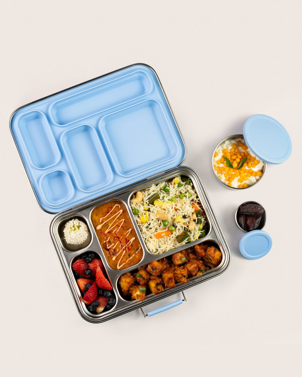 Stainless-steel leakproof bento lunchbox in blue packed with Indian meal including vegetable biryani, paneer tikka, mixed berries, and additional snack containers filled with boondi raita and dates, perfect for kids' school lunches.