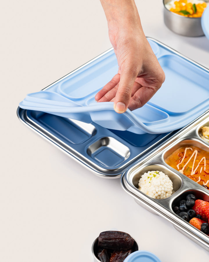 Leakproof blue stainless-steel lunchbox with a silicone lid being removed, showing compartments filled with Indian meal including paneer tikka, sabudana, mixed berries, and dates, perfect for kids' school lunches.