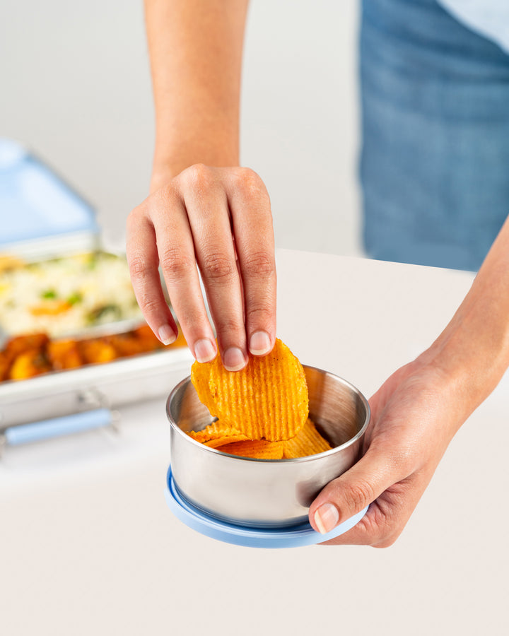 Hand holding a stainless steel snack container with blue silicone lid, filled with crispy potato chips, next to a bento lunch box with various dishes, perfect for keeping snacks fresh in India