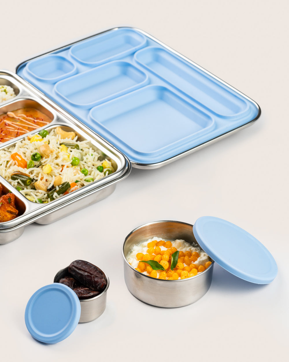 Stainless steel snack container set with blue silicone lids, featuring a large and small container filled with dates and a mixed snack, next to a bento lunch box with various dishes, perfect for use in India