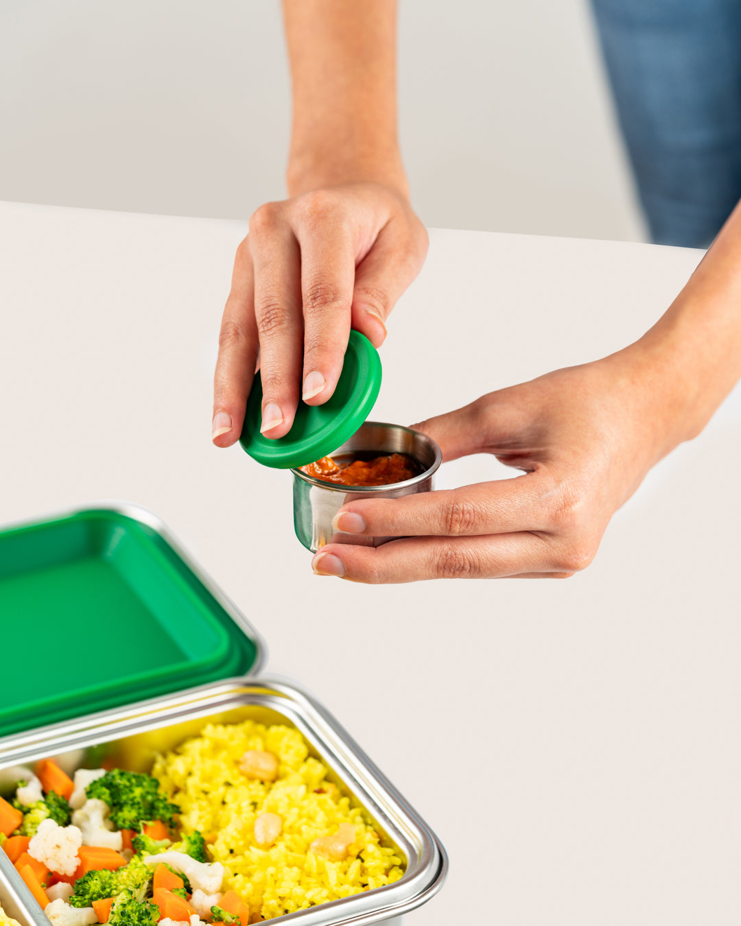 Hand sealing a small stainless steel container with green silicone lid filled with chutney, next to a lunch box with rice and vegetables, perfect for Indian snacks