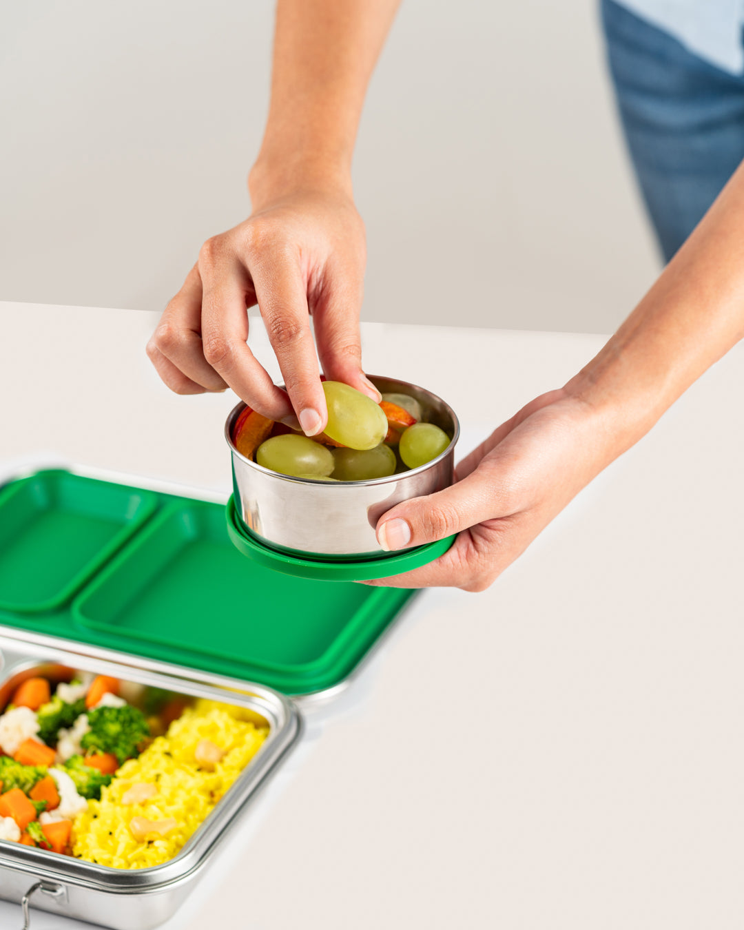 Hand holding stainless steel snack container with green silicone lid, filled with grapes and carrots, next to a lunch box with healthy food, ideal for Indian snacks