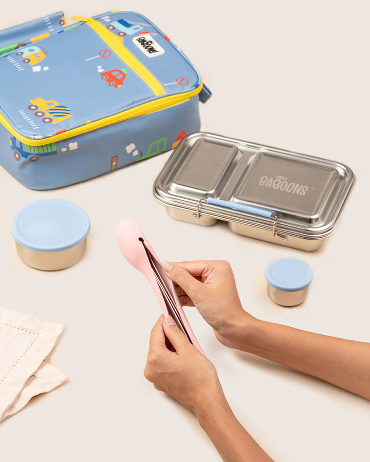 Bento box set with colorful lunch bag snack containers and hand holding silicone case spoon fork and knife set in soft pink on a light surface