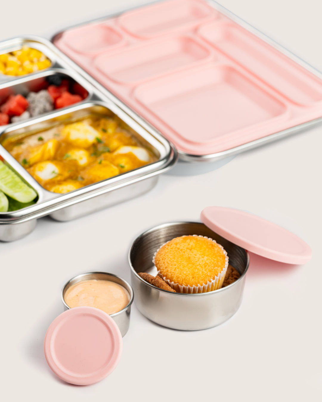 Stainless steel snack containers with pink silicone lids holding a cupcake and dip, perfect for bento lunch boxes in India