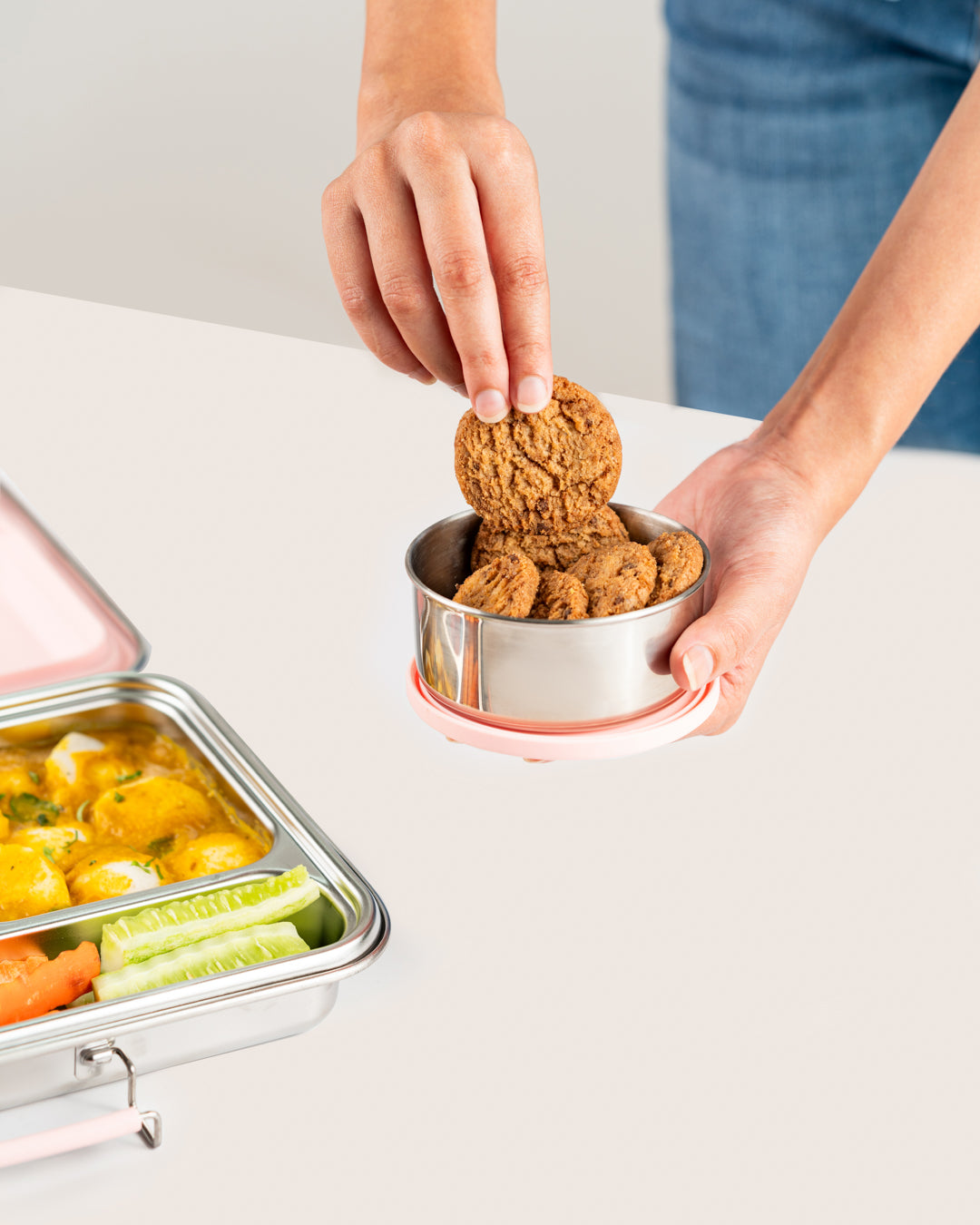 Person placing cookies into a large stainless steel snack container with a pink lid next to a lunch box with vegetables and curry, showcasing a snack container set for lunch boxes in India.